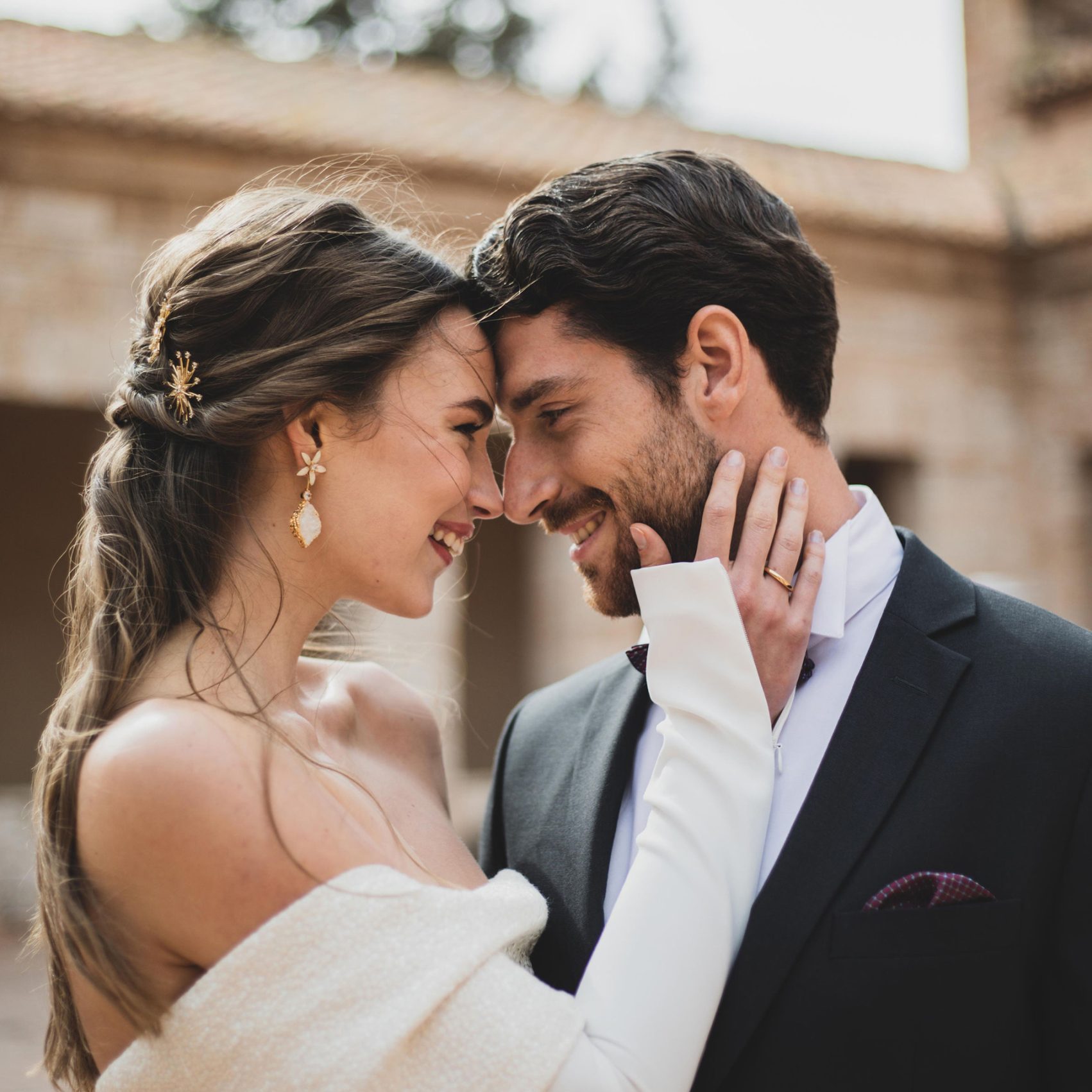 Pareja de novios abrazándose y acercando sus frentes, mostrando un momento íntimo y romántico al aire libre, con la novia usando un vestido sin mangas y un velo largo