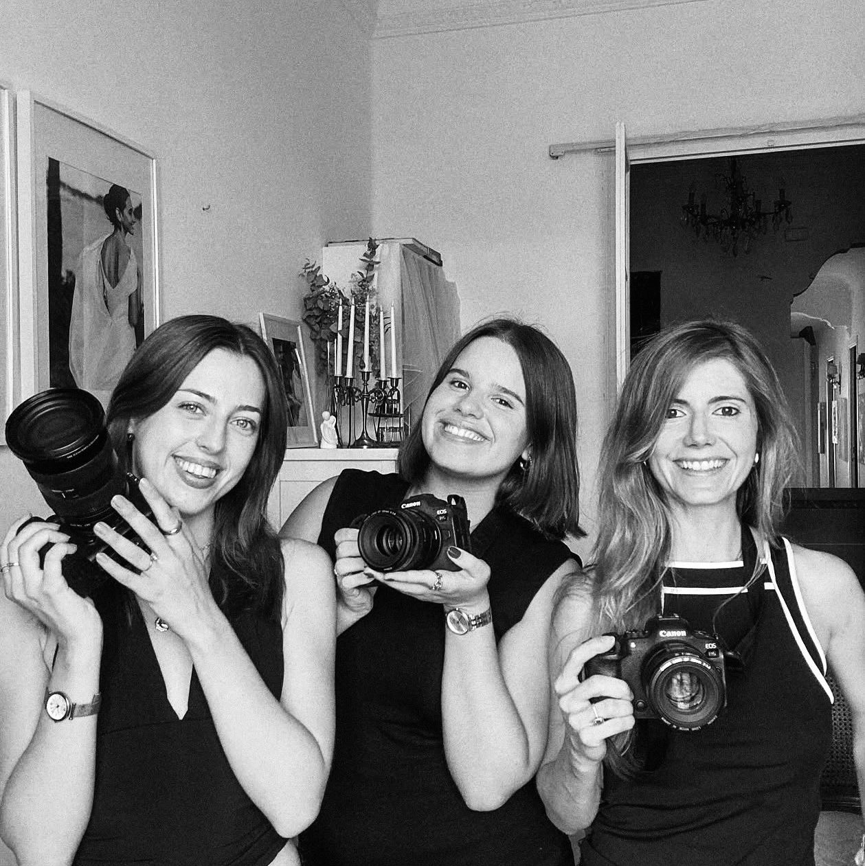 Mujeres sonrientes posando juntas con cámaras fotográficas, en blanco y negro, mostrando su equipo y actitud profesional.