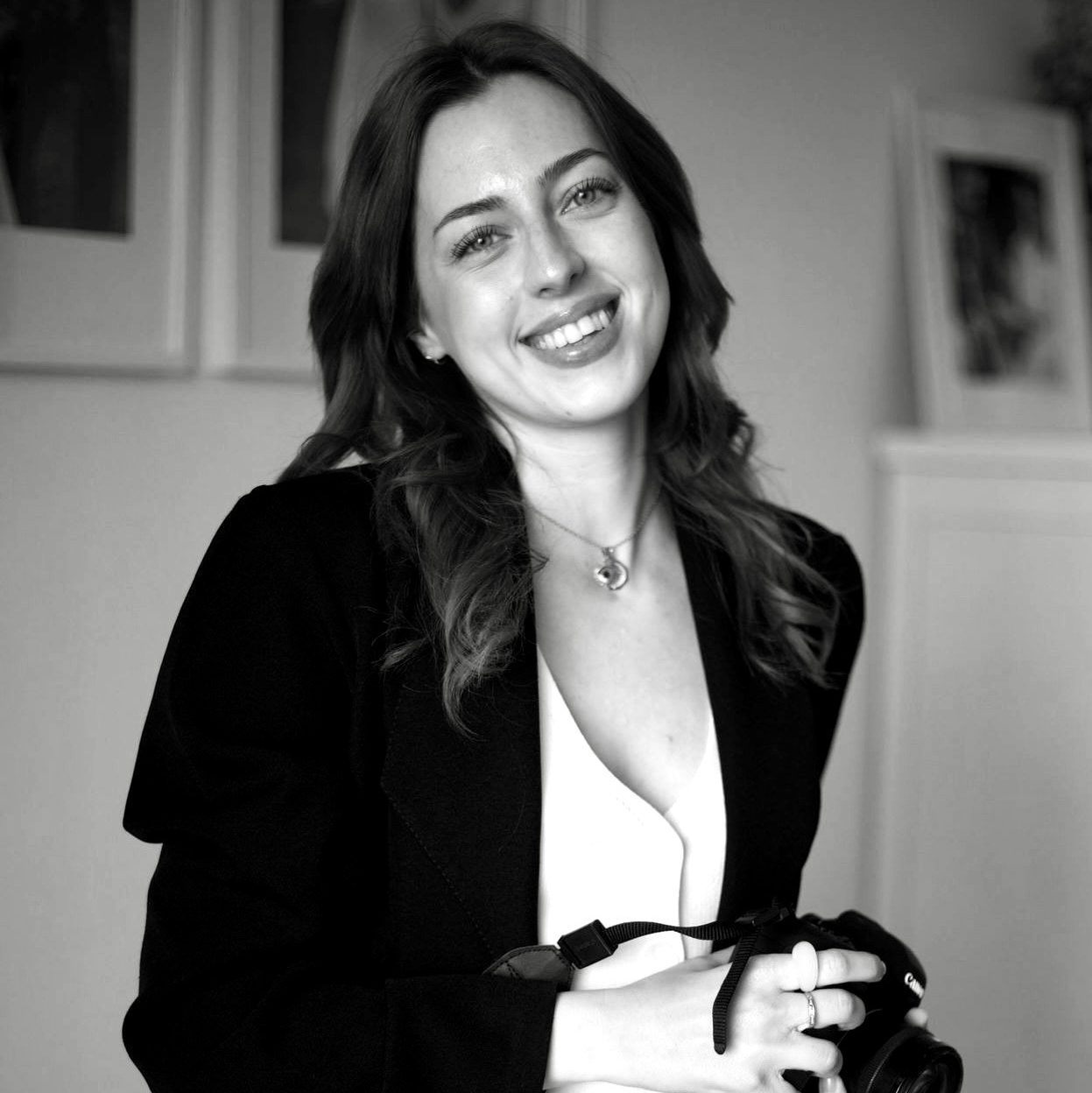 Mujer sonriendo mientras sostiene una cámara fotográfica, en blanco y negro, con un fondo de decoración minimalista